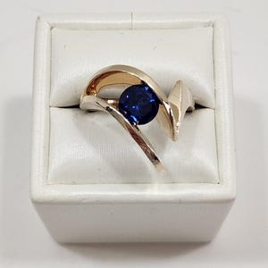 May include: A gold ring featuring a single round, deep blue gemstone set in the center. The band has a smooth, polished finish with a curved, asymmetrical design that wraps around the stone, creating an open, flowing shape on both sides.