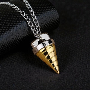 Könnte beinhalten: Ein goldfarbener Anhänger in Form einer Eistüte hängt an einer silbernen Kette. Der Anhänger hat eine silberne Oberseite und eine goldene Tüte mit spiralförmigen Rillen. Die Halskette wird vor einem dunklen Hintergrund präsentiert.