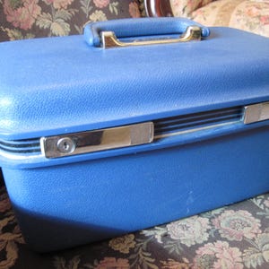 May include: A vintage blue hard-sided suitcase with a gold-colored handle and silver-toned latches. The suitcase has a textured surface and is rectangular in shape. A classic travel accessory.