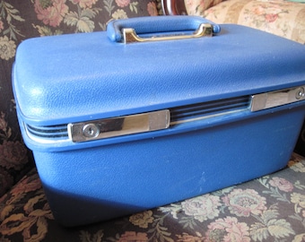 May include: A vintage blue hard-sided suitcase with a gold-colored handle and silver-toned latches. The suitcase has a textured surface and is rectangular in shape. A classic travel accessory.