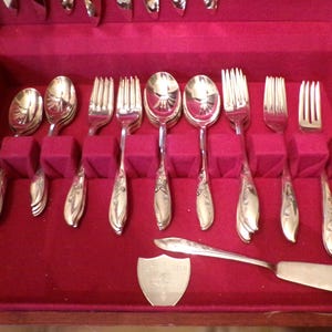 May include: A set of silver-colored flatware, including spoons, forks, and serving utensils, arranged in a red velvet-lined case. The silverware has ornate floral detailing on the handles. The case is open, displaying the neatly organized pieces.