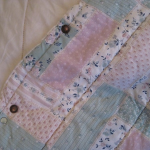 May include: Close-up of a patchwork quilt with a floral pattern in pastel colors. The quilt features squares of pink, blue, and white fabric, with floral and dotted designs. Two brown buttons are visible on the quilt's edge.