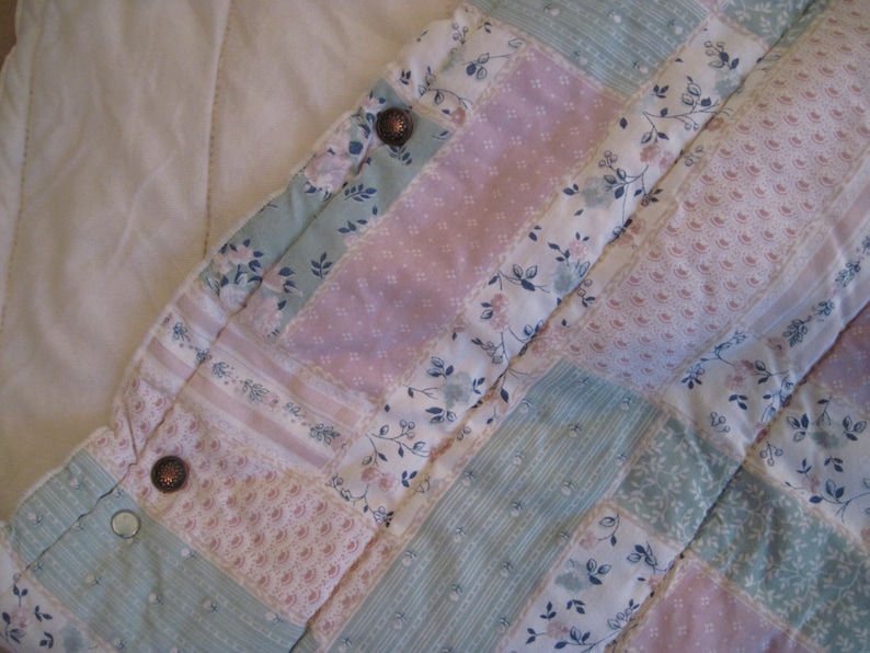 May include: Close-up of a patchwork quilt with a floral pattern in pastel colors. The quilt features squares of pink, blue, and white fabric, with floral and dotted designs. Two brown buttons are visible on the quilt's edge.