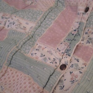 May include: Close-up of a quilted fabric with a patchwork design. The quilt features rectangular patches in pastel pink, mint green, and cream, adorned with floral and dotted patterns. Dark brown buttons are visible along the seam.