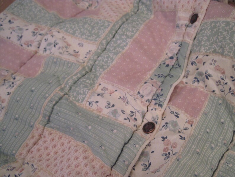 May include: Close-up of a quilted fabric with a patchwork design. The quilt features rectangular patches in pastel pink, mint green, and cream, adorned with floral and dotted patterns. Dark brown buttons are visible along the seam.