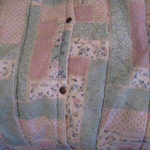 May include: Close-up of a patchwork quilt with a buttoned closure. The quilt features a mix of floral and solid patterns in pastel pink, green, and white. The buttons are a dark brown color. The quilt is likely handmade.