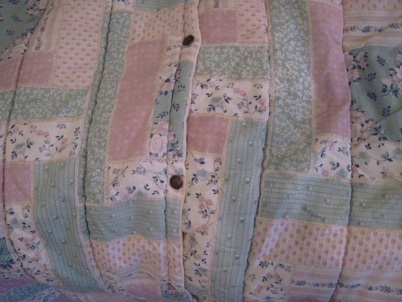 May include: Close-up of a patchwork quilt with a buttoned closure. The quilt features a mix of floral and solid patterns in pastel pink, green, and white. The buttons are a dark brown color. The quilt is likely handmade.