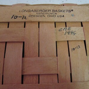 Pu&ograve; includere: Primo piano di un cestino Longaberger tessuto a mano. Il cestino &egrave; fatto di strisce di legno marrone chiaro intrecciate tra loro. Il testo "LONGABERGER BASKETS HANDWOVEN DRESDEN, OHIO USA" &egrave; stampato in alto. I numeri "10-12" e "CAE 1994" sono scritti a mano.