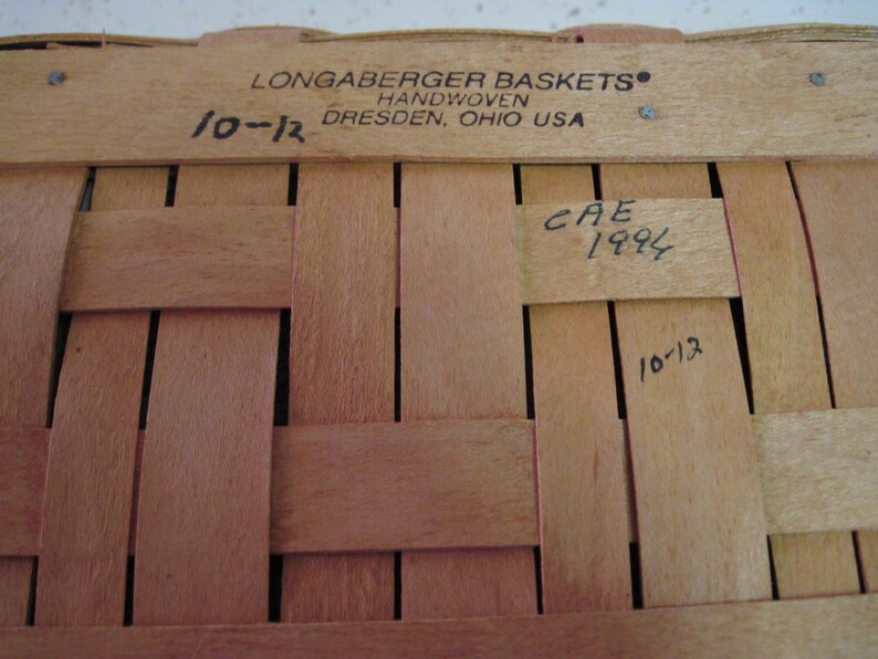 Pu&ograve; includere: Primo piano di un cestino Longaberger tessuto a mano. Il cestino &egrave; fatto di strisce di legno marrone chiaro intrecciate tra loro. Il testo "LONGABERGER BASKETS HANDWOVEN DRESDEN, OHIO USA" &egrave; stampato in alto. I numeri "10-12" e "CAE 1994" sono scritti a mano.