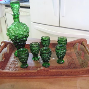 Puede incluir: Un juego de bar vintage de vidrio verde con un diseño de racimo de uvas, que incluye una jarra y seis vasos pequeños a juego. El conjunto se exhibe en una bandeja de madera tallada con asas.