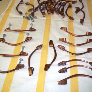 May include: Various copper-toned cabinet handles and pulls, some with screws, displayed on a yellow and white striped cloth. The hardware features both curved and angular designs, ideal for furniture renovation or new installations.