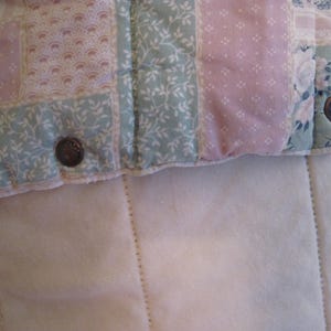 May include: Close-up of a quilted fabric with a patchwork design. The quilt features squares of pastel pink and green fabric with floral and dotted patterns. Two decorative buttons are visible on the edge of the quilt, which is resting on a white, textured surface.