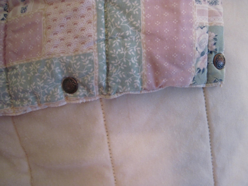 May include: Close-up of a quilted fabric with a patchwork design. The quilt features squares of pastel pink and green fabric with floral and dotted patterns. Two decorative buttons are visible on the edge of the quilt, which is resting on a white, textured surface.