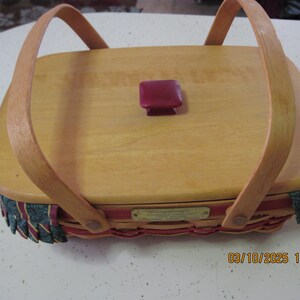 Pu&ograve; includere: Cesto in legno chiaro di forma rettangolare con coperchio e due manici ad arco. Il coperchio presenta un piccolo pomello quadrato bordeaux. I bordi del cesto sono decorati con un bordo intrecciato rosso, verde e oro.
