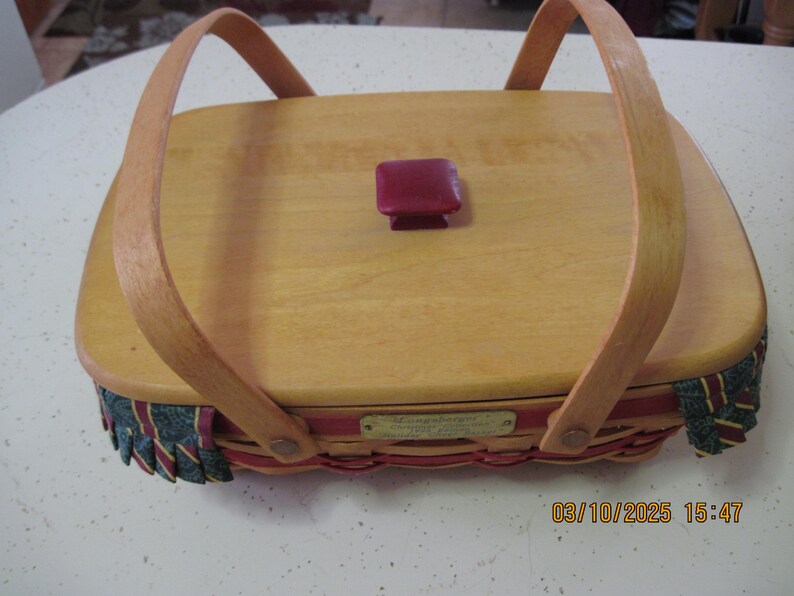 Pu&ograve; includere: Cesto in legno chiaro di forma rettangolare con coperchio e due manici ad arco. Il coperchio presenta un piccolo pomello quadrato bordeaux. I bordi del cesto sono decorati con un bordo intrecciato rosso, verde e oro.