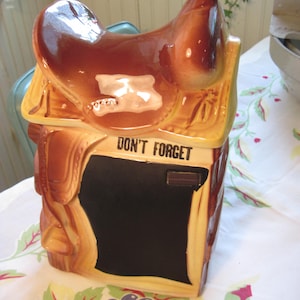 May include: A brown ceramic cookie jar shaped like a saddle, with a black chalkboard area and the words "DON'T FORGET" printed on it. The jar has a glossy finish and is set on a floral tablecloth.