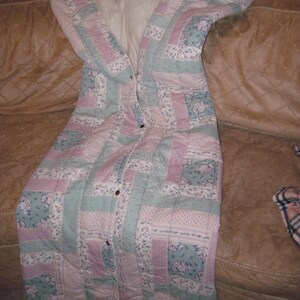 May include: A quilted sleeping bag in pastel colors of pink, blue, and white, with a floral pattern. The sleeping bag is buttoned up and appears to be made of soft fabric. The sleeping bag is on a brown couch.