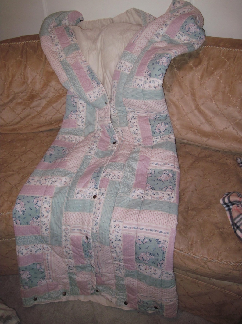 May include: A quilted sleeping bag in pastel colors of pink, blue, and white, with a floral pattern. The sleeping bag is buttoned up and appears to be made of soft fabric. The sleeping bag is on a brown couch.