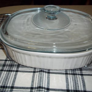 May include: An oval, white casserole dish with a clear glass lid and metal handles. The lid has a circular design in the center. The dish sits on a black and white plaid cloth. A kitchenware item.