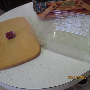 Pu&ograve; includere: Un tagliere in legno chiaro con un manico rosso scuro &egrave; posizionato accanto a un contenitore di plastica rettangolare trasparente. Un cestino decorativo con un motivo rosso, verde e blu &egrave; sullo sfondo. Gli oggetti sono su una superficie bianca maculata.