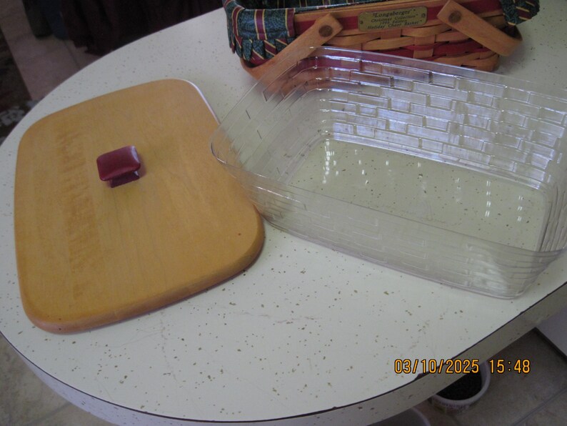 Pu&ograve; includere: Un tagliere in legno chiaro con un manico rosso scuro &egrave; posizionato accanto a un contenitore di plastica rettangolare trasparente. Un cestino decorativo con un motivo rosso, verde e blu &egrave; sullo sfondo. Gli oggetti sono su una superficie bianca maculata.