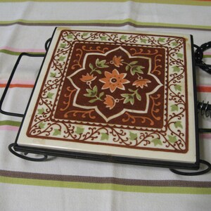 May include: A vintage ceramic hot pad with a brown, orange, and green floral design. The pad is set in a black metal frame with a handle and a spring.