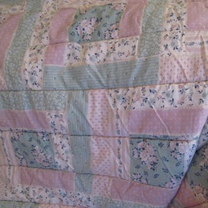 May include: A quilted blanket with a patchwork design. The quilt features alternating stripes of light pink, pale blue, and floral patterned fabric. The floral patterns include white and blue flowers. The quilt is folded, showing its texture and construction.