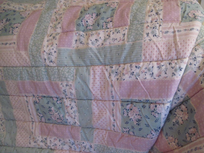 May include: A quilted blanket with a patchwork design. The quilt features alternating stripes of light pink, pale blue, and floral patterned fabric. The floral patterns include white and blue flowers. The quilt is folded, showing its texture and construction.