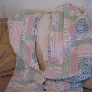 May include: A quilted jacket in a patchwork design, featuring pastel pink and mint green floral and dotted patterns. The jacket is folded over a light-colored pillow, showcasing its interior lining and button closure.