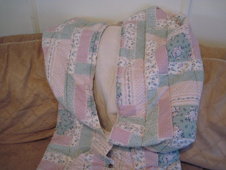May include: A quilted jacket in a patchwork design, featuring pastel pink and mint green floral and dotted patterns. The jacket is folded over a light-colored pillow, showcasing its interior lining and button closure.