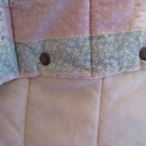 May include: Close-up of a quilted fabric with floral and geometric patterns in pastel pink, blue, and white. The quilt has a soft, textured appearance with visible stitching and metal button closures. The overall design is reminiscent of vintage or cottagecore aesthetics.
