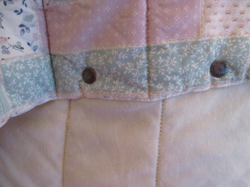May include: Close-up of a quilted fabric with floral and geometric patterns in pastel pink, blue, and white. The quilt has a soft, textured appearance with visible stitching and metal button closures. The overall design is reminiscent of vintage or cottagecore aesthetics.