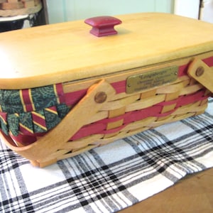 Pu&ograve; includere: Un cestino da picnic rettangolare in legno con coperchio in legno chiaro e pomello rosso. Il cestino &egrave; intrecciato con legno chiaro e dettagli rossi e presenta una fodera in tessuto a fantasia verde e rossa sui bordi. Una placca metallica &egrave; visibile sul lato.