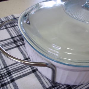 May include: A white casserole dish with a clear glass lid and a silver handle. The dish has a blue trim and is sitting on a black and white checkered cloth. The lid has a decorative pattern.
