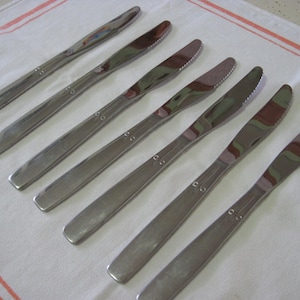 May include: A set of six silver butter knives with smooth, rounded handles. The knives are arranged in a row on a white and orange striped surface.