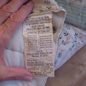 May include: A close-up of a quilted fabric with a tag attached. The tag reads "RESIN TREATED POLYESTER FIBER BATTING" and lists the size as 60 x 77 inches. The fabric has floral and geometric patterns in soft colors.