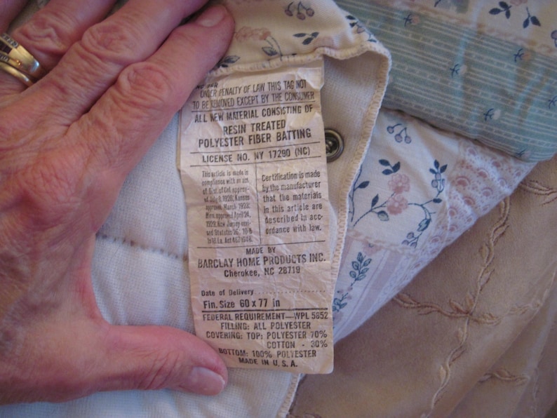 May include: A close-up of a quilted fabric with a tag attached. The tag reads "RESIN TREATED POLYESTER FIBER BATTING" and lists the size as 60 x 77 inches. The fabric has floral and geometric patterns in soft colors.