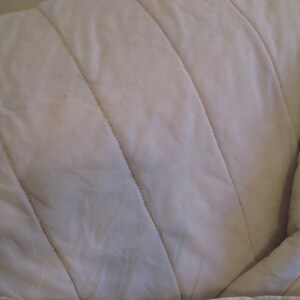 May include: Close-up of a cream-colored, quilted fabric. The fabric has a soft, textured appearance with diagonal stitching creating a series of parallel lines. The overall look is simple and neutral, suitable for bedding or upholstery.