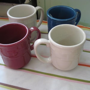 May include: Four ceramic mugs in shades of white, blue, and burgundy are arranged on a striped surface. Each mug has a handle and a textured design. The mugs are suitable for hot beverages.