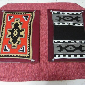 May include: Two decorative ceramic tiles with rug designs. One tile features a red background with a black and gold geometric pattern. The other tile has a black and gray geometric pattern on a white background. Both tiles have a burgundy border.