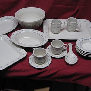 以下が含まれることがあります： 青い花のアクセントが付いた白いセラミック食器のコレクション。セットには、サービングボウル、プレート、カップ、ソーサー、サービングスプーン、マグカップが含まれます。赤いテーブルクロスの上に配置されています。