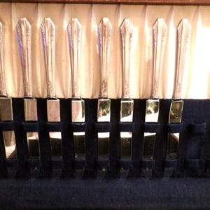 May include: A set of silver knives in a wooden case. The knives are arranged in a row, with their blades facing down. The case has a blue velvet lining and a cream-colored fabric background.
