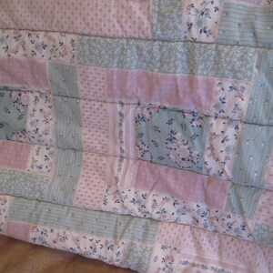 May include: A patchwork quilt featuring a mix of floral and solid fabric blocks in pastel pink, blue, and white. The quilt has a quilted texture with visible stitching, creating a cozy and handmade aesthetic. The overall design is a mix of floral patterns and solid colors.