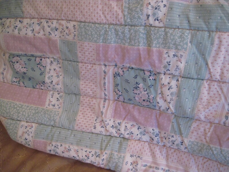 May include: A patchwork quilt featuring a mix of floral and solid fabric blocks in pastel pink, blue, and white. The quilt has a quilted texture with visible stitching, creating a cozy and handmade aesthetic. The overall design is a mix of floral patterns and solid colors.