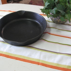 May include: A black cast iron frying pan with a long handle rests on a white and striped kitchen towel. The towel has orange, green, and brown stripes. A decorative bowl with green leaves is in the background.