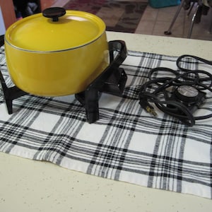 Puede incluir: Una olla de fondue eléctrica amarilla vintage con asa y pomo negros. La olla se encuentra sobre un soporte negro, y un cable de alimentación negro con un dial de control de temperatura está al lado. La olla está sobre un mantel a cuadros blanco y negro.
