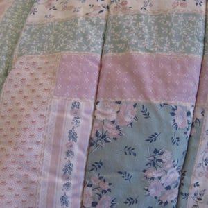 May include: Close-up of a patchwork quilt featuring a mix of floral and patterned fabrics in pastel colors. The quilt has sections of pink, light green, and white with floral and lace designs. The quilt is a cozy bedding item.