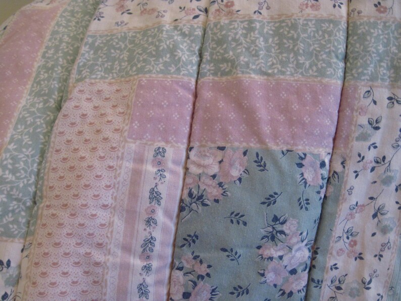 May include: Close-up of a patchwork quilt featuring a mix of floral and patterned fabrics in pastel colors. The quilt has sections of pink, light green, and white with floral and lace designs. The quilt is a cozy bedding item.
