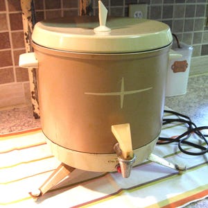May include: Vintage beige and cream-colored electric coffee percolator. The appliance has a cross design on the front and a spigot. It sits on three legs and has a cream-colored lid and handle. A power cord is attached.