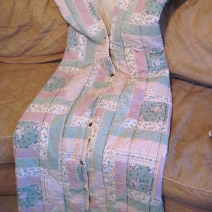 May include: A quilted robe in pastel colors of pink, green, and white, with a floral pattern. The robe has a button-up front and a shawl collar. The robe is lying on a brown couch.